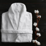 Clean folded bathrobe and branch of cotton on black wooden background, flat lay